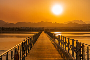 Fototapeta na wymiar Picturesque bright orange sunset over silhouette of desert mountains of Egypt and coastline with reflection in sea water and wooden bridge road leading to sunlight on summer evening at vacation