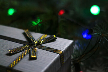 Defocus gray gift with close-up gold bow ribbon on pine or fir tree blurred background with glowing festive green, red and blue light bulb at night. Christmas present. Holiday box. Out of focus