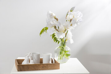A bouquet of three white irises and a fern in a transparent vase on the table. Two ceramic tea in the wooden tray. Breakfast