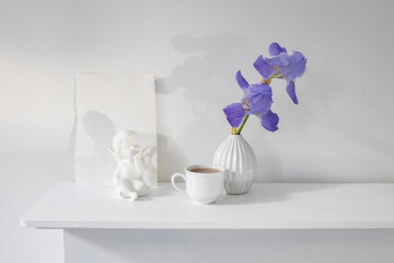 Blue iris in a corrugated vase with a piece of craft paper on a white table. Figurine of the angel. Cup of tea. Place for text