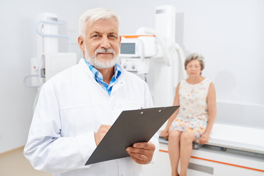 Front view of grey haired radiologist smiling, writing history of illness after ultrasound exams of female patient. Blurred background with oldest woman in dress waiting near ultra sound equipment.