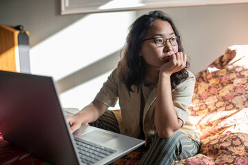 Young distracted female in casualwear and eyeglasses sitting in front of laptop