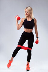 A beautiful girl with a sports figure performs exercises with an elastic band for fitness and dumbbells.