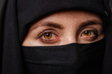 Close up portrait of young, adult woman in black burqa  with hidden face, isolated on black...
