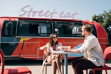 Young couple interacting while having fast food in outdoor cafe against street truck - Powered by Adobe