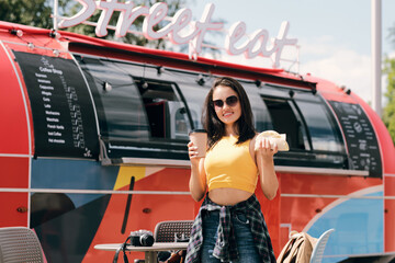 Happy young brunette woman holding glass of coffee and hotdog against street food truck