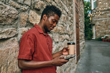 Happy young African man with drink texting in smartphone while standing by stone wall