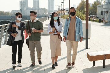 Young Asian colleagues in protective masks taking walk in park on sunny day