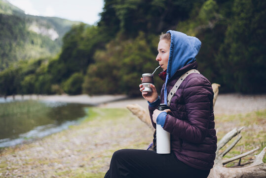 Young adult caucasian woman drinking traditional argentinian yerba mate, sitting in a trunk while contemplates the lake on a cold morning. Patagonia landscape in Argentina. Copyspace