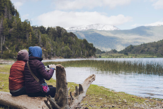 Back view of a young adult couple sharing traditional argentinian yerba mate, sitting in a trunk while contemplates the lake on a cold morning. Patagonia landscape in Argentina.