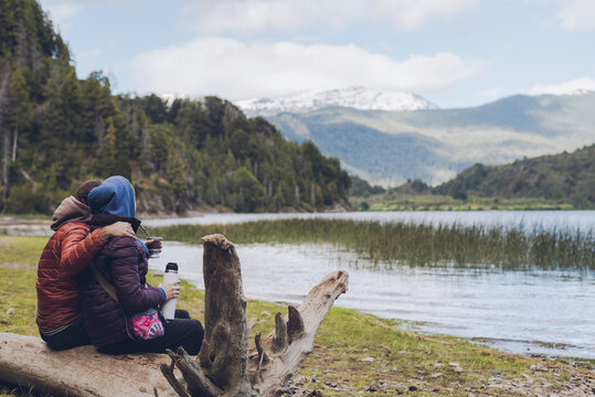 Back view of a young adult couple sharing traditional argentinian yerba mate, sitting in a trunk while contemplates the lake on a cold morning. Patagonia landscape in Argentina.