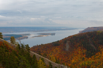 Autumn view of the opposite shore across a wide river and an island in the middle. A river with an island on the background of a mountain forest with bright multicolored foliage and large gray clouds