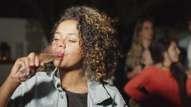 African-American woman drinking alcohol and looking at camera. Medium shot of happy female person holding glass of champagne, posing for camera during night party with friends. Party, emotion concept