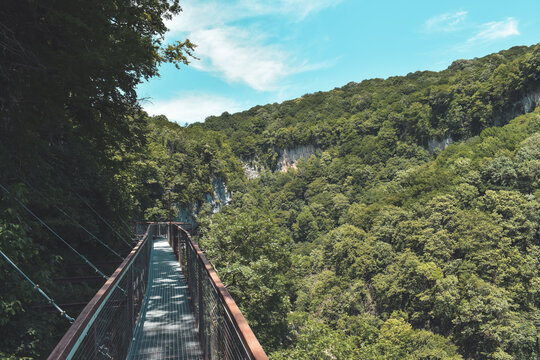 Suspension bridge in Okatse Canyon, village Gordi, Imereti region, Georgia. Narrow path surrounded by hills covered by green trees. Blue sky with light clouds above. Georgian tourist attraction