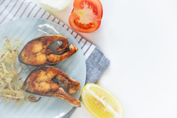 Fried fish with onions lies on a plate, on a waffle towel, next to a lemon and a tomato on a white background.