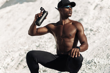 muscular African American athletic man resting after workout, holding bottle of water, in mountains outdoors