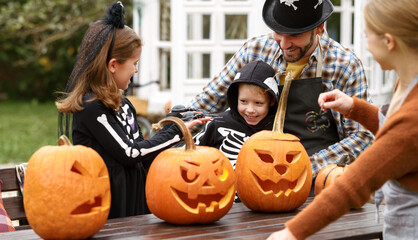 Happy young family in Halloween costumes carving pumpkins together in backyard