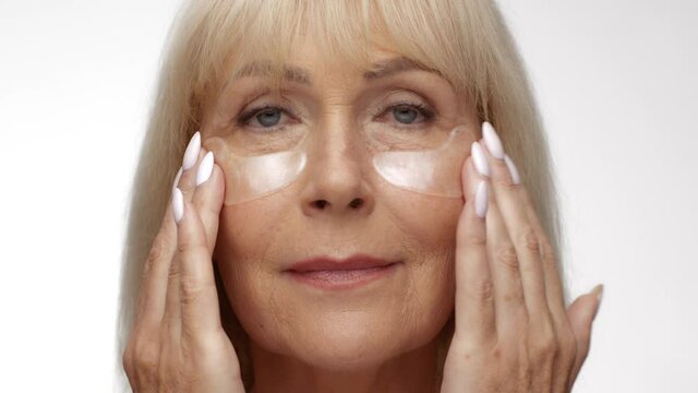 Anti wrinkles skin care. Senior blonde woman applying collagen gel patches under her eyes skin against crow's feet