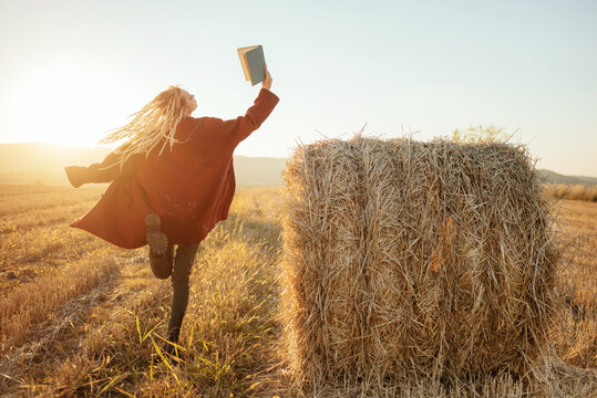 Cheerful young woman, dancing and jumping while reading an exciting book in the field outside
