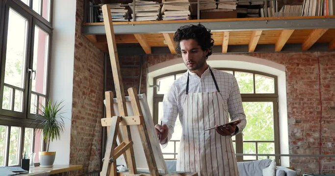 Attractive Hispanic painter engrossed in painting pictures on easel, guy wear apron holds palette, paintbrush makes brushstrokes on canvas. Creative hobby, professional occupation, art studio concept