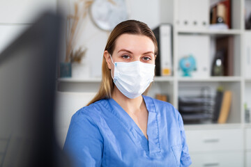 Fototapeta na wymiar Portrait of young female doctor in face mask working on laptop in clinic office