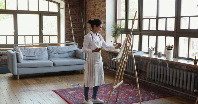 In spacious light loft workshop room Indian woman painter creates masterpieces of art hold palette and paintbrush, drawing on canvas. Talented person, creative hobby, professional occupation concept