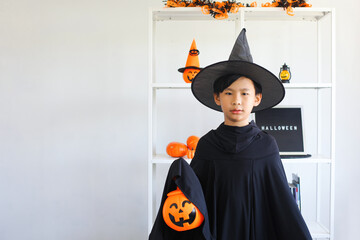Cute Asian boy celebrating Halloween by wearing witch costume and holding Halloween decoration...