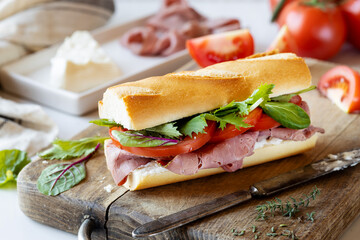 Delicious baguette sandwich with roast beef, soft cheese, tomatoes and green salad leaves on a wooden cutting board