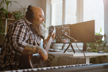 Mature composer plays bas guitar recording track at workplace in home studio
