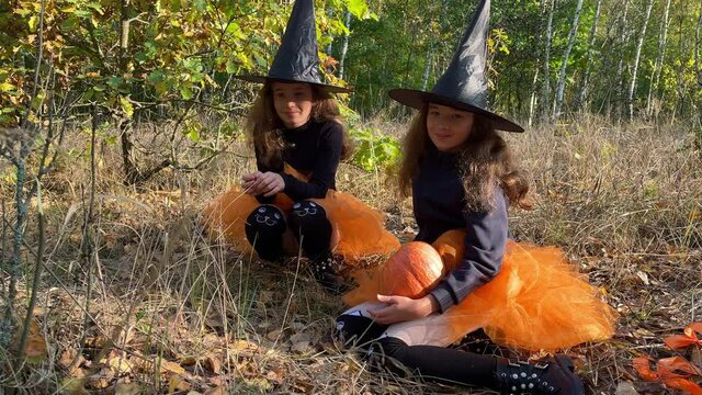 Twin girls dressed like witches are seating in a forest and laughing.