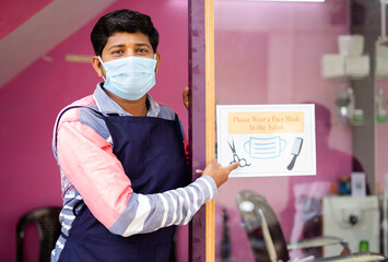 Barber with medical mask showing wear mask at saloon sign board to avoid coronavirus or covid-19...