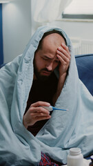 Ill person wrapped in blanket looking at thermometer for temperature measurement at home. Man with...