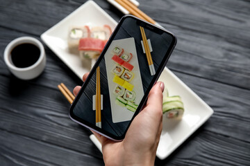 Woman holding mobile phone and taking picture of plate with set of delicious sushi rolls on grey wooden table