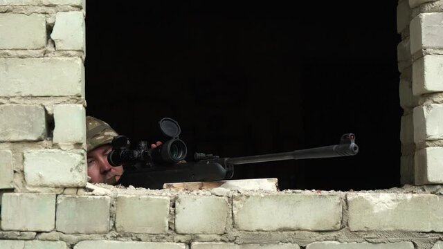 a soldier in camouflage takes aim from the window of an abandoned building with a sniper rifle