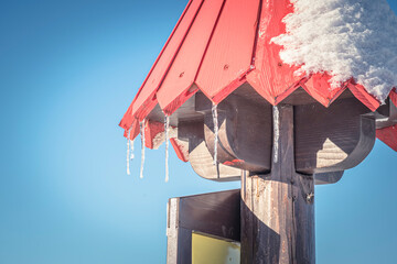 Long icicles on a wooden mountain sign, Slovakia. Red paint on pointy roof, dark natural pole made of wood. Selective focus on texture, blurred background.