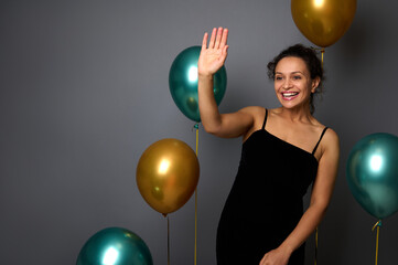Fototapeta premium Beautiful woman dressed in elegant black evening dress stands against a gray wall background with golden and green metallic inflated balloons, waves hello with her hand, smiles with toothy smile