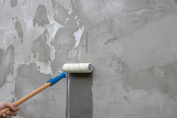 A worker applies the primer to the wall with a long-handled roller. A man's hand rolls a primer onto a gray wall. Wet footprint on a plastered wall. House facade renovation concept.