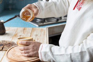 Ethnic linen shirt on the man. A man with a beard in the kitchen pours honey into a cup. Cooking dessert in a rustic kitchen.