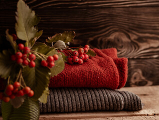 a bouquet of red berries in a glass vase, warm knitted things are lying on a wooden table on a dark background