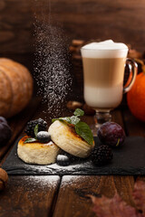Two cheesecakes and a glass mug with latte stand on a wooden table on a black stand. Powdered sugar is poured on top of the cheesecakes. Decorated with berries and mint