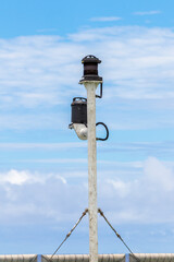 Ship's stern light post with two navigation lights against cloudy blue sky. Turnbuckles and steel wires are holding the pole upright.