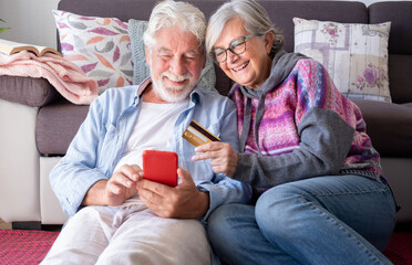 Happy senior couple in living room at home having fun shopping online paying  with credit card, e-commerce, buyer, consumerism concept