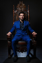 Elegant man in a classic blue suit sitting in a carved wooden chair against a dark background.