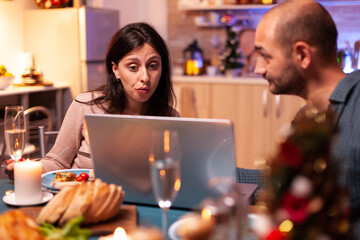 Joyful couple browsing on social media reading entertainment message while sitting at dining table in xmas decorated kitchen. Happy family celebrating christmas holiday enjoying winter season together