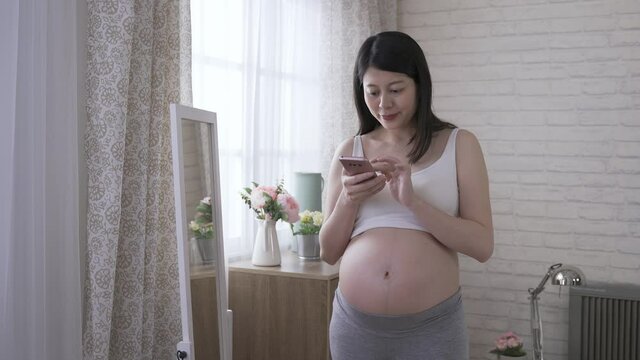expectant mother is tucking her hair and touching her tummy gently while using a smartphone to shoot her own image in the mirror with joy at home.