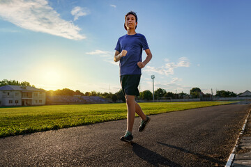 concept of sports and health - teen boy runs along the stadium track, a soccer field with green grass.