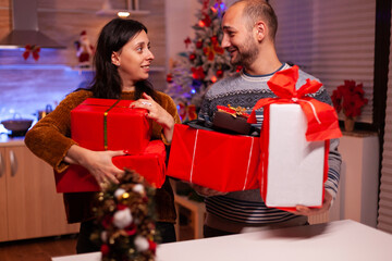 Obraz na płótnie Canvas Happy married couple holding secret present gift with ribbon on it during christmas holiday standing in xmas decorated kitchen. Smiling family enjoying winter season celebrating christmastime together
