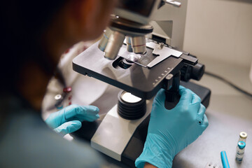 Lab assistant in latex gloves looks into microscope at sample in laboratory closeup