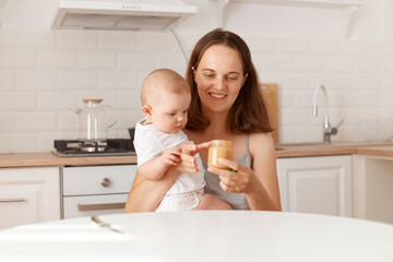 Portrait of happy young adult dark haired female sitting in light kitchen, feeds her little daughter with fruit or vegetable puree, opens jar, healthy feeding up.