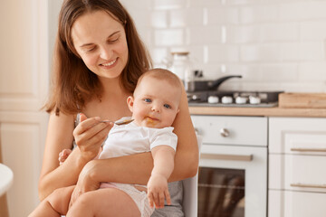 Smiling happy young adult dark haired female feeding her little daughter with fruit or vegetable puree, sitting in light kitchen, expressing care and love.